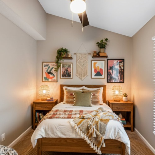 A brightly lit, modern boho teenager's bedroom featuring a solid wooden twin bed flanked by lit nightstands, neutral bedding with a tasseled throw, a woven rug, potted plants, and simplified gallery wall decor with a large round mirror and framed prints.