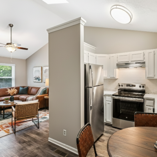 A bright, open-plan interior showcases a modern living room with a leather sectional and a patterned rug flowing into a white kitchen area featuring stainless steel appliances and a dark wood breakfast table