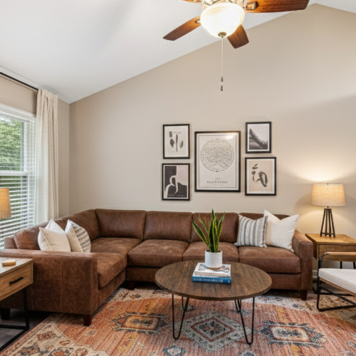 A brightly lit, modern living room with a brown leather sectional sofa, a colorful patterned area rug, two wooden end tables with lamps, a woven accent chair, and a gallery wall of black-framed abstract art under a vaulted ceiling with a fan.