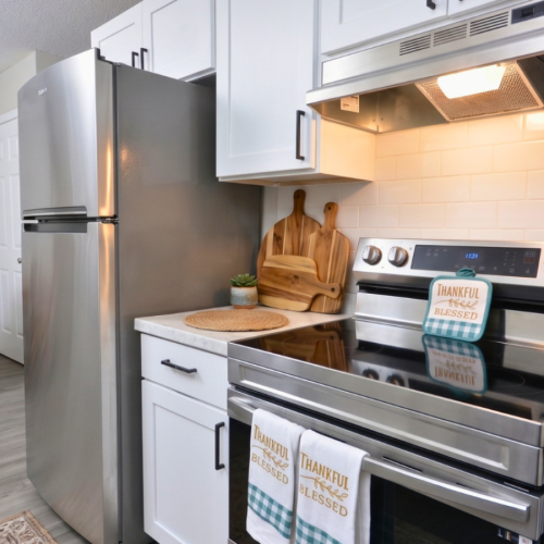 Renovated kitchen at Sherrill Place Apartments featuring stainless steel appliances, white shaker-style cabinetry, black hardware, subway tile backsplash, and marble-inspired countertops.
